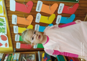 Dziewczynka w odświętnej sukience pozuje na tle zawieszonych na ścianie kolorowych, świątecznych skarpet podpisanych imionami dzieci.